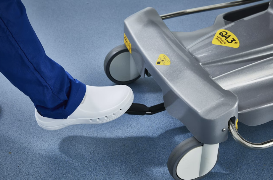 Person in blue scrubs pressing a foot pedal on medical equipment with wheels.
